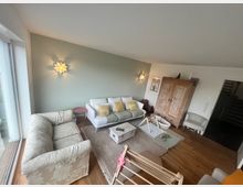 Wohnzimmer mit Holzboden, zwei Sofas und einem Sessel; eine grüne Wand mit zwei blumenförmigen Wandlampen; ein großer Holzschrank sowie ein Kinder-Spielbereich mit Teppich und Spielbogen; große Fensterfront auf der linken Seite.