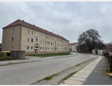 Wohnblock in einer ruhigen Wohngegend mit beigefarbener Fassade und rotem Ziegeldach. Vor dem Gebäude führt eine gepflasterte Straße entlang, daneben Gehwege, Rasenflächen und ein großer, blattloser Baum.