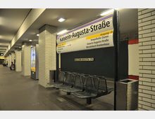U-Bahnhof mit weiß gefliesten Säulen, einer Beschilderung "Kaiserin-Augusta-Straße", grauen Sitzbänken, und Wegweisern zu verschiedenen Ausgängen. Im Hintergrund ist ein Bahnsteig mit dunkler Wand und vereinzelte Personen zu sehen.