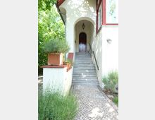 Eingangsbereich eines Hauses mit weißer Fassade, einem bogenförmigen Torbogen und einer roten Holztür am Ende einer Steintreppe. Links und rechts der Treppe befinden sich Geländer, sowie Pflanzen und eine Terrasse mit Pflastersteinen im Vordergrund.