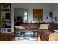 Wohnzimmer mit Ecksofa aus braunem Stoff, dekoriert mit hellen Kissen. Im Vordergrund ein Glastisch mit Zeitschriften, im Hintergrund ein Bücherregal, ein Gemälde an der Wand und ein Durchgang zu einem anderen Raum mit sichtbarem Fenster.