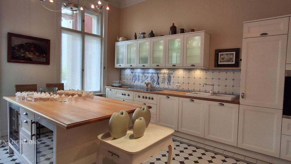 Helle, klassische Küche mit weißen Einbauschränken, Holz-Arbeitsflächen und einer langen Kücheninsel; an der Rückwand ein blau-weiß gefliestes Muster mit dekorativem Motiv. Große Fenster bringen viel Tageslicht, und der Boden ist in einem schwarz-weißen geometrischen Fliesenmuster gehalten.