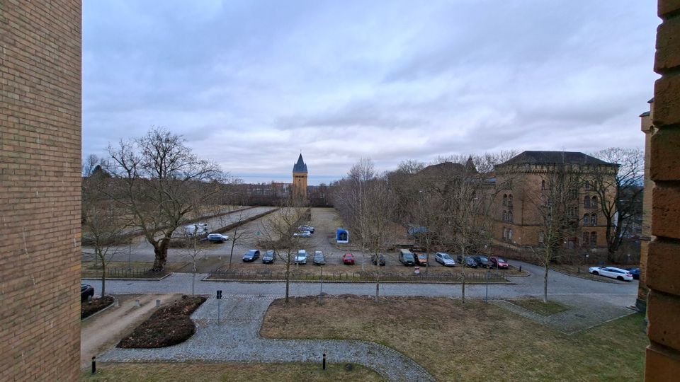 Blick von einem Backsteingebäude auf ein Gelände mit gepflasterten Wegen, einer Wiese und einem Parkplatz mit mehreren Autos. Im Hintergrund stehen laublose Bäume, ein hoher Turm mit spitzem Dach und ein großes historisches Backsteingebäude unter bewölktem Himmel.