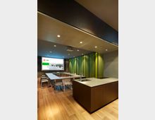 Moderner Konferenzraum mit Holzfußboden, einem U-förmigen Tisch mit weißen Stühlen und einer großen Präsentationsfläche an der Wand. Im Vordergrund befindet sich eine Kücheninsel mit Spüle, im Hintergrund grün akzentuierte Wände.