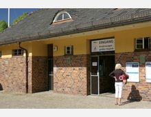 Backstein- und gelb verputztes Gebäude mit Schieferdach und Dachfenster, das als Eingang für Kinderwagen und großes Gepäck dient. Eine Frau steht auf einem gepflasterten Weg und liest Schilder neben der Eingangstür.