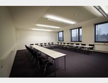 Ein moderner Besprechungsraum mit rechteckiger Tischanordnung in U-Form, umgeben von schwarzen Stühlen. Der Raum hat beige Wände, großen Fenstern mit Blick nach draußen und deckenmontierte Leuchtstofflampen.