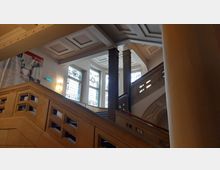 Innenansicht eines historischen Treppenhauses mit dekorativen Glasfenstern, Geländern aus Stein mit rechteckigen Aussparungen und verzierten Säulen. Die Decke ist mit geometrischen Vertiefungen gestaltet, und die Szene wird von natürlichem Licht durch die Fenster beleuchtet.