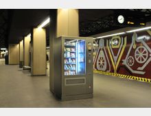 Ein U-Bahn-Bahnhof mit modernem Design, beigefarbenen Säulen und einer Wand mit geometrischen Mustern in Rot, Gelb und Grau. Im Vordergrund steht ein beleuchteter Snackautomat, im Hintergrund eine Uhr und ein Informationsdisplay.