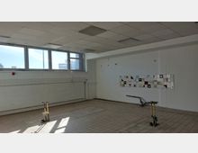 Ein leerer Raum mit Fliesenboden, weißen Wänden und einer großen Fensterfront an der oberen Wandseite. An einer Wand befindet sich eine farbige Kachelverzierung mit geometrischen Mustern, und im Raum sind Rohre und Installationen sichtbar.