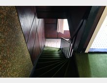 Holzgetäfeltes Treppenhaus mit grüner Teppichtreppe, schwarzem Geländer und floral gemusterter Tapete an einer Seite; im Hintergrund ein Fenster mit Vorhang.