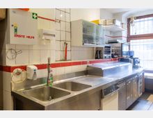 Kleine Gastro-Küche mit Edelstahl-Arbeitsfläche, Doppelspülbecken und integrierten Geräten wie einem Toaster und einer Kühlvitrine. An der weiß-gekachelten Wand befinden sich ein Erste-Hilfe-Kasten, ein Papierspender sowie offene Regale und Schränke.