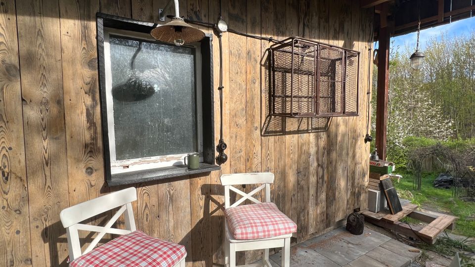 Überdachte Veranda an einer Holzhütte mit Bretterwand, kleinem Fenster, Wandlampen und einem angebrachten Drahtregal. Davor stehen zwei weiße Barhocker mit karierten Sitzkissen auf Steinplatten; rechts öffnet sich der Blick ins grüne Gelände.