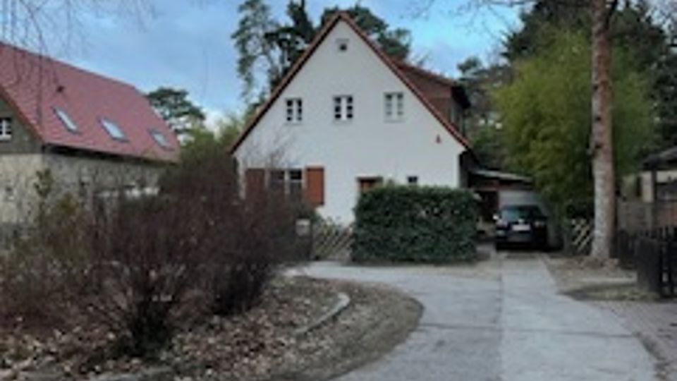 Einfahrt aus Beton zu einem kleinen, weiß verputzten Wohnhaus mit steilem Giebeldach und braunen Fensterläden. Links verläuft ein geschwungener Weg mit niedrigen Sträuchern, rechts stehen ein Zaun und ein Carport neben hohen, teils kahlen Bäumen.