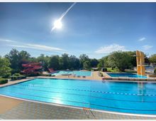 Ein Freibad mit mehreren Schwimmbecken, darunter ein großes Becken mit Schwimmbahnen im Vordergrund. Im Hintergrund sind eine rote Wasserrutsche, ein Sprungturm und grüne Bäume sichtbar.
