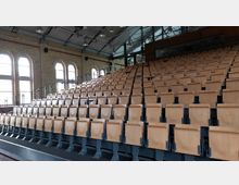 Holzbestuhlte Sitzreihen in einem Hörsaal oder Veranstaltungsraum mit Backsteinmauerwerk und großen Fensterbögen; das Dach ist mit Stahlträgern und Industrieleuchten ausgestattet.