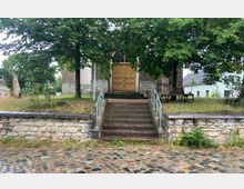 Treppenaufgang zu einem Gebäude mit gelber Doppeltür aus Holz, flankiert von Bäumen. Im Vordergrund Kopfsteinpflaster und eine niedrige Mauer aus hellen Steinen.