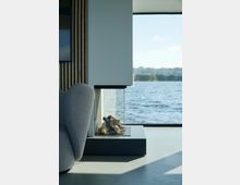 Modernes Wohnzimmer mit einem Glas-Kamin in der Ecke, auf dem Holzscheite dekorativ angeordnet sind. Durch ein großes Panoramafenster bietet sich ein Blick auf einen See und eine bewaldete Uferlandschaft.