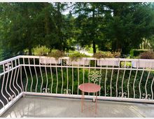 Balkon mit weißem, verziertem Geländer und darauf befestigten Blumenkästen, gefüllt mit grünen und blühenden Pflanzen. Im Hintergrund ist eine dichte Baumreihe mit hohen Nadelbäumen und einer Grünfläche sichtbar, während auf dem Balkon ein kleiner runder Tisch steht.