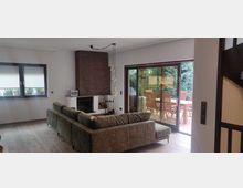 Wohnzimmer mit einem modernen Kamin aus roten Ziegelsteinen, einer grünen Eckcouch und einer großen Glasschiebetür, die zu einer Terrasse mit Holztisch und Stühlen führt. Im Raum befinden sich helle Wände, Holzfußboden und eine Fensterfront mit geschlossenen Rollläden links.