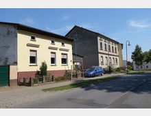 Straßenansicht mit zwei Wohnhäusern, eines mit gelber Fassade und Vorgarten, das andere mit grauer, leicht verwitterter Fassade. Ein blauer Pkw parkt vor dem grauen Gebäude, daneben eine Laterne und einige Bäume.