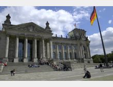 Das Bild zeigt die Vorderseite des Reichstagsgebäudes in Berlin, einem historischen Parlamentsgebäude mit einer Fassade aus Säulen und dem Schriftzug „Dem Deutschen Volke“. Im Vordergrund befinden sich Menschen auf den Treppen und auf dem Platz, während eine deutsche Fahne an einem Fahnenmast im Wind weht.