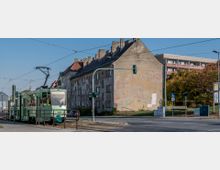 Straßenszene mit einer grünen Straßenbahn an einem Haltepunkt, daneben ein altes, teils verputztes Mehrfamilienhaus mit sichtbaren Schäden an der Fassade. Im Hintergrund stehen moderne Wohngebäude und Bäume mit herbstlichem Laub.