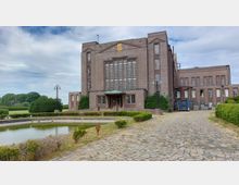 Backsteingebäude mit symmetrischer Fassade und großen vertikalen Fenstern, flankiert von kleineren Anbauten. Im Vordergrund ein gepflasterter Weg, ein ovaler Teich und gepflegte Büsche, im Hintergrund eine weitläufige Landschaft mit Bäumen.