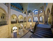 Innenansicht eines historischen Gebäudes mit doppelt geschwungenen Treppen, Rundbögen aus hellem Stein und verzierten Geländern. Die Kuppeldecke ist blau mit einem Muster aus dekorativen Sternen, und große Fenster lassen Tageslicht herein.