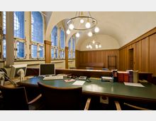 Ein historischer Innenraum mit gewölbter Decke, großen mehrteiligen Fenstern mit Sprossen und Holztäfelung an den Wänden. Ein Schreibtisch im Vordergrund ist mit Büchern, Monitoren und Schreibutensilien ausgestattet; an der Decke hängen zwei runde Kronleuchter.