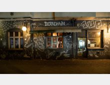 Außenansicht einer Bar mit dem Namen "Berndhain" in einer dunklen Straße, deren Fassade mit weißer, skizzenhafter Graffiti-Kunst wie Ketten, Totenköpfen und Flammen verziert ist. Durch ein erleuchtetes Fenster ist das Innere der Bar zu sehen, während eine alte Markise und Straßenlaternen den Eingangsbereich beleuchten.