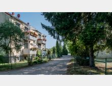 Mehrfamilienhaus mit Balkonen und Blumenkästen, gelegen an einem schmalen, asphaltierten Weg, umgeben von hohen Nadelbäumen und einem Maschendrahtzaun. Im Hintergrund sind weitere grüne Büsche und Bäume zu sehen.