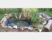 Ein kleiner, künstlicher Teich mit einer schwarzen Kunststoffwanne, umgeben von Natursteinen, hohen Gräsern und Holzstämmen, in einer Gartenlandschaft mit grünem Hintergrund.