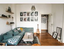 Wohnzimmer mit einer Eckcouch in hellblaugrau, dekoriert mit gemusterten Kissen. Ein rustikaler Couchtisch aus Holz mit Rollen steht auf einem gemusterten Teppich, an der Wand hängen mehrere schwarz-weiße Bilderrahmen. Im Raum stehen eine Gitarre und an der rechten Wand lehnen gerahmte Bilder auf dem Holzboden.