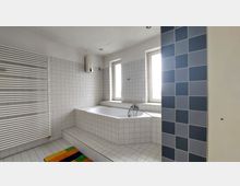 Helles Badezimmer mit einer erhöhten Badewanne, weiß gefliesten Wänden und einem blau gefliesten Bereich. Zwei Fenster sorgen für Tageslicht, und ein weißer Handtuchheizkörper ist an der Wand befestigt.