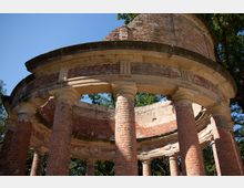 Teilansicht einer historischen, runden Ruinenstruktur aus rotem Backstein mit offenen Bögen und Säulen. Im Hintergrund sind Bäume und ein blauer Himmel zu sehen.