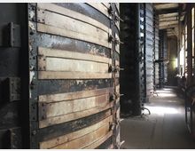 Industrieller Innenraum mit einer Reihe großer, horizontal geschichteter Metalltanks, die verrostet und verwittert aussehen. Der Boden besteht aus Betonplatten, und im Hintergrund fällt Licht durch ein Fenster auf die Maschinen.