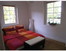 Schlafzimmer mit einem Doppelbett, das eine bunte Bettwäsche in Rot-, Orange- und Gelbtönen hat. Zwei große Fenster mit weißen Rahmen lassen Tageslicht herein; eines hat einen Blumenkasten mit Pflanzen. Der Raum hat hell gestrichene Wände, einen Holzboden und eine schmale weiße Stehlampe in der Ecke.