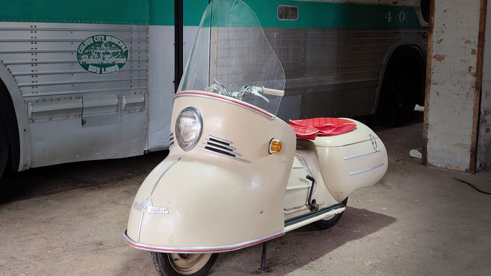 Innenraum einer alten Garage oder eines Depots mit Betonboden und gemauertem Pfeiler, in dem ein cremefarbener Vintage-Roller mit Windschutzscheibe und rotem Sitz steht. Im Hintergrund ist die Seitenwand eines grün-silbernen Busses mit Nieten und Fenstern zu sehen.