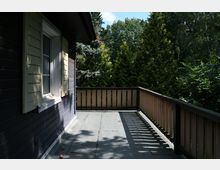 Blick auf eine Terrasse mit grauem Bodenbelag und einem hölzernen Geländer, das die Fläche umschließt. Links ist eine Hauswand mit dunkelbrauner Holzverkleidung und einem Fenster mit cremefarbenen Fensterläden zu sehen, im Hintergrund dichte grüne Bäume.