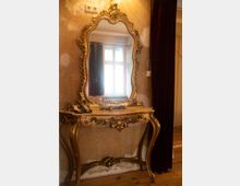 Vergoldeter Barock-Spiegel mit geschwungenem Rahmen, montiert über einem reich verzierten Konsolentisch in einem Raum mit abgetragenen Wänden und Holzboden; im Spiegel ist ein Fenster mit Vorhängen sichtbar.