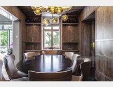 Esszimmer mit elegantem Holzdekor, rundem dunklen Esstisch und gepolsterten Lederstühlen. Im Hintergrund befinden sich ein großes Fenster mit Blick ins Grüne sowie Wandregale mit dekorativen Objekten, ergänzt durch einen auffälligen Kronleuchter.