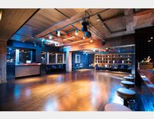 Ein moderner Veranstaltungsraum mit offener Tanzfläche, Holzboden und Betonwänden. Die Decke verfügt über sichtbare Metallträger und warme, neonartige Beleuchtung. Im Hintergrund stehen Sitzbereiche mit Regalen, die dekorative Objekte enthalten. Rechts im Vordergrund ist eine Theke mit Barhockern sichtbar.