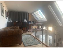 Dachgeschoss-Wohnzimmer mit schrägen Decken und großen Dachfenstern, die viel Licht einlassen. Die Einrichtung umfasst ein braunes Ledersofa, einen Teppich, eine kleine Bar mit hohen Stühlen sowie ein Treppengeländer aus Glas und Metall.