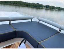 Ein Ausschnitt einer Bootsterrasse mit gepolsterten blauen Sitzbänken und gelben Nähten, umgeben von einem Geländer. Im Hintergrund ist ein ruhiger See mit bewaldetem Ufer und vereinzelten Gebäuden zu sehen.