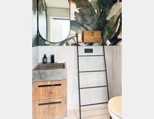 Modernes Badezimmer mit einer grauen Steinwaschbeckenplatte, einem Unterschrank aus Holz, einer schwarzmetallenen Leiter als Handtuchhalter, und tropisch bedruckter Tapete mit großen Blättern. Im Hintergrund ist eine helle Wand mit Steckdosen sichtbar.