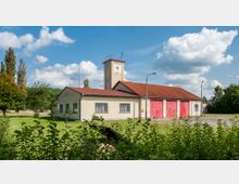 Ein einstöckiges Feuerwehrgebäude mit beigem Putz, roten Garagentoren und einem Turm mit Uhr, umgeben von einer Rasenfläche und Bäumen, unter einem sonnigen Himmel mit einzelnen Wolken.