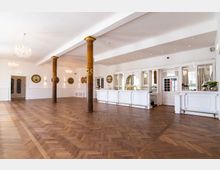 Großer, eleganter Veranstaltungsraum mit hölzernem Fischgrätboden, verzierten Holzsäulen, weiß gestrichenen Wänden und einer weißen Bar mit Spiegeln im Hintergrund. Mehrere Kronleuchter hängen von der Decke, und runde Wanddekorationen schmücken die Wände.