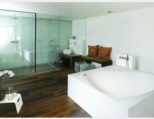 Modernes Badezimmer mit freistehender, weißer Badewanne im Vordergrund und einer großen, ebenerdigen Dusche mit Glaswänden im Hintergrund. Der Raum hat einen dunklen Holzfußboden, eine weiße Wand, eine Sitzbank mit bunten Kissen und dekorative Elemente wie Orchideen auf einem kleinen Tisch.