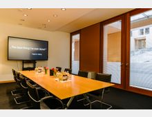 Konferenzraum mit einem rechteckigen Tisch aus Holz, umgeben von schwarzen Lederstühlen. Im Hintergrund befindet sich ein großer Bildschirm mit einem Zitat, sowie bodentiefe Fenster mit Holzrahmen und Jalousien, die einen Blick nach draußen ermöglichen.