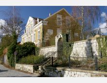 Gelbes, zweistöckiges historisches Gebäude mit weißen Fensterrahmen und dekorativen Elementen, umgeben von einer Steinmauer mit einem Torbogen, der mit Efeu bewachsen ist. Im Vordergrund führt eine Treppe zu einem umzäunten Garten, während Bäume und Sträucher die Umgebung prägen.